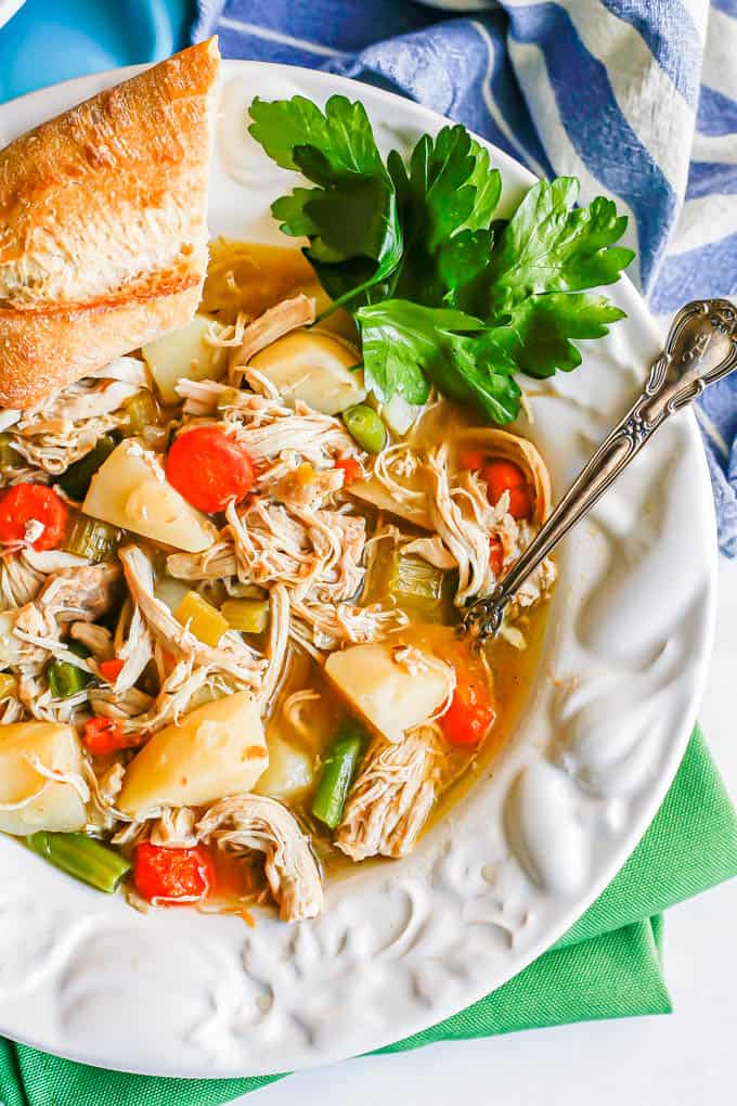 A white bowl with chunky chicken stew with parsley and a hunk of bread and a spoon resting in the side