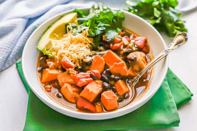 A low white bowl of sweet potato and black bean chili with toppings and a spoon on the side