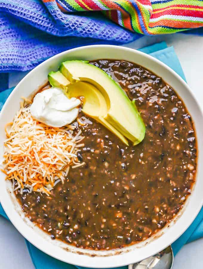 A low white bowl with black bean soup topped with shredded cheese, sour cream and avocado