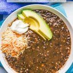 A low white bowl with black bean soup topped with shredded cheese, sour cream and avocado and a text overlay on the photo