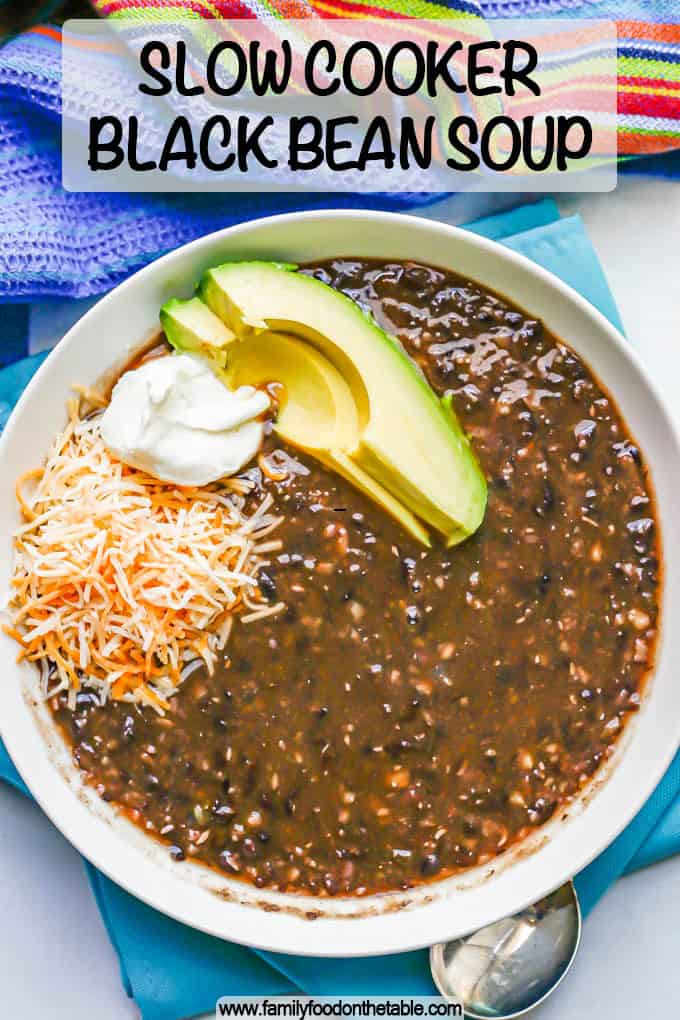 A low white bowl with black bean soup topped with shredded cheese, sour cream and avocado and a text overlay on the photo