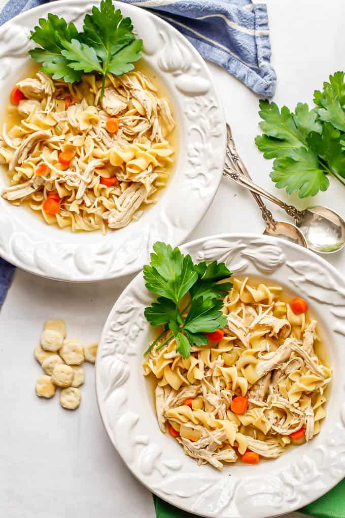 Two white soup bowls with chicken noodle soup garnished with fresh parsley with oyster crackers and spoons to the side