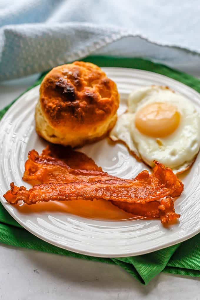 A small white plate with strips of bacon, a fried egg and a biscuit