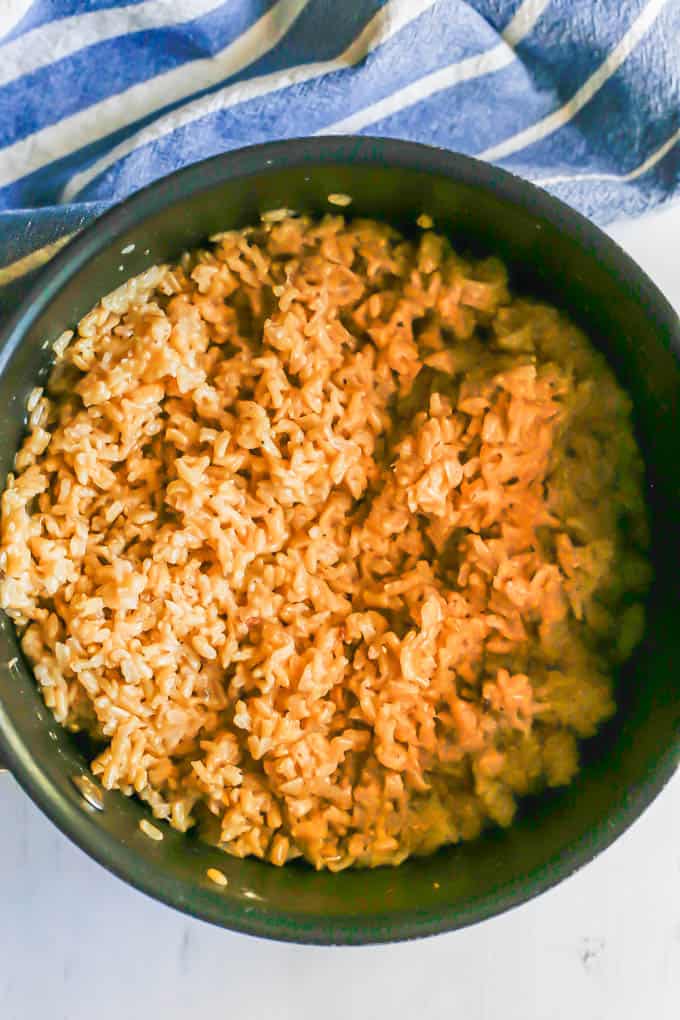 Brown rice cooked with onions and broth in a medium dark pan with a blue striped towel in the background