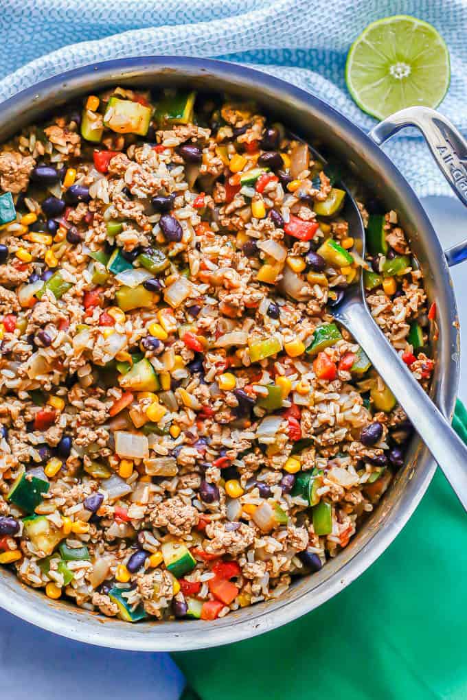 A metal serving spoon resting in a large skillet with ground turkey, rice, veggies, beans and corn with green and turquoise napkins to the side and a halved lime