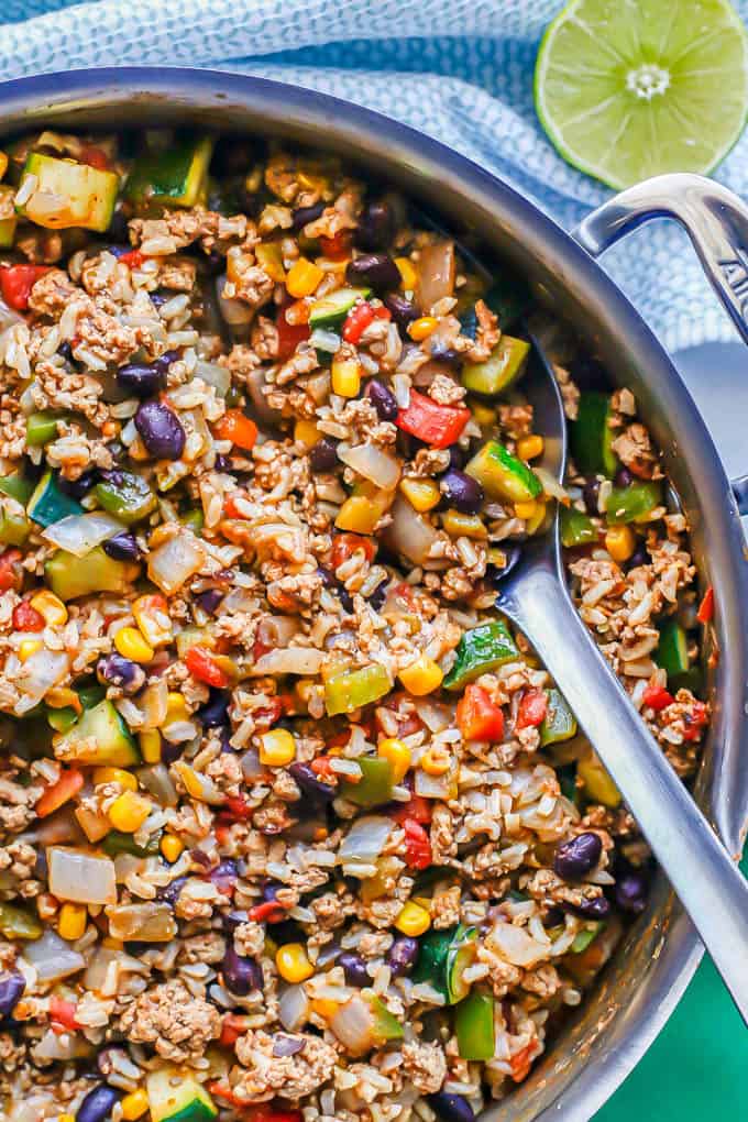 Close up of a metal serving spoon resting in a large skillet with ground turkey, rice, veggies, beans and corn