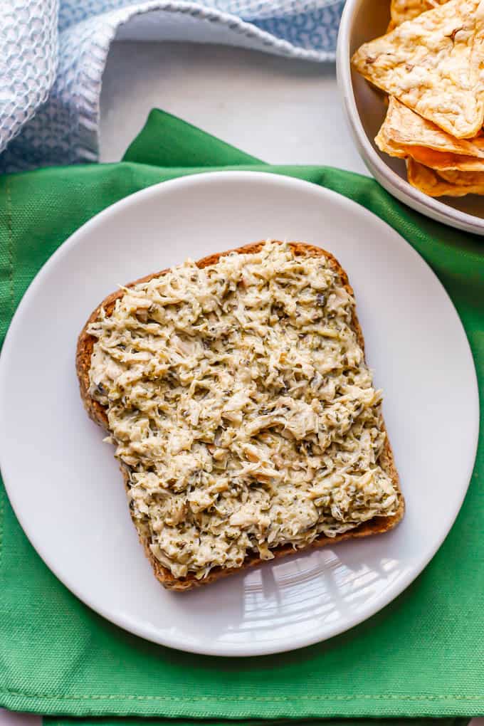 A small white plate with a piece of toast covered with tuna salad and a small bowl of chips nearby