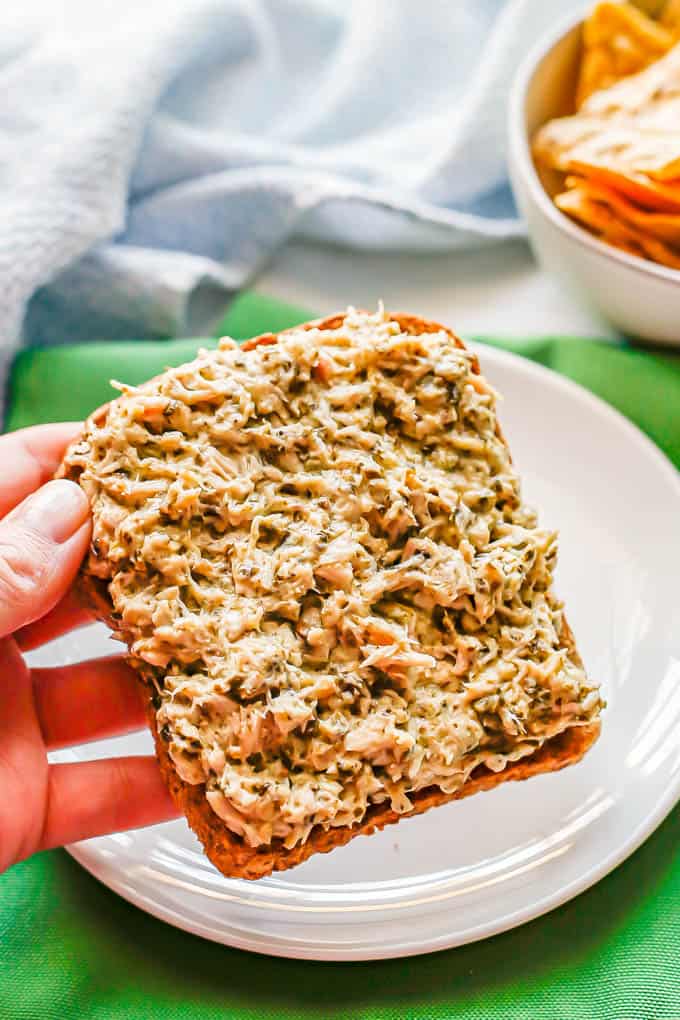 A hand picking up a piece of pesto tuna salad toast from a white plate