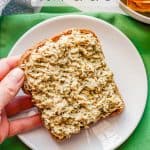 A hand holding a piece of toast with pesto tuna salad spread on it with a text overlay on the photo