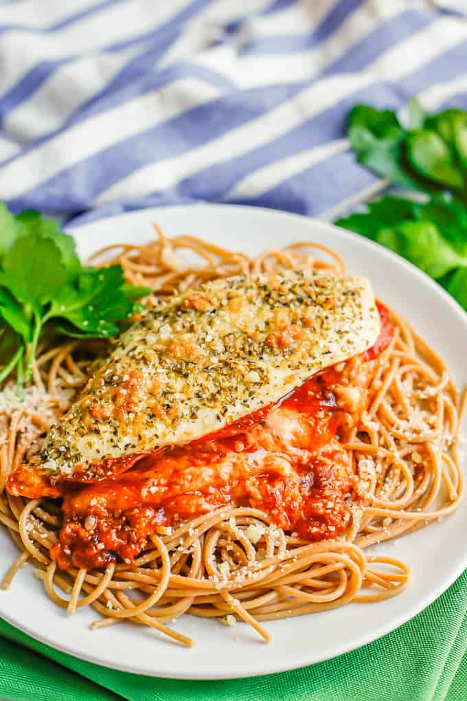 A white plate with spaghetti topped with a pizza stuffed chicken breast and sprigs of parsley to the side for garnish