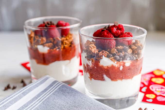 Two yogurt parfaits in clear glasses with layers of yogurt, strawberry sauce, granola, berries and chocolate chips and Valentine's cards in the background.