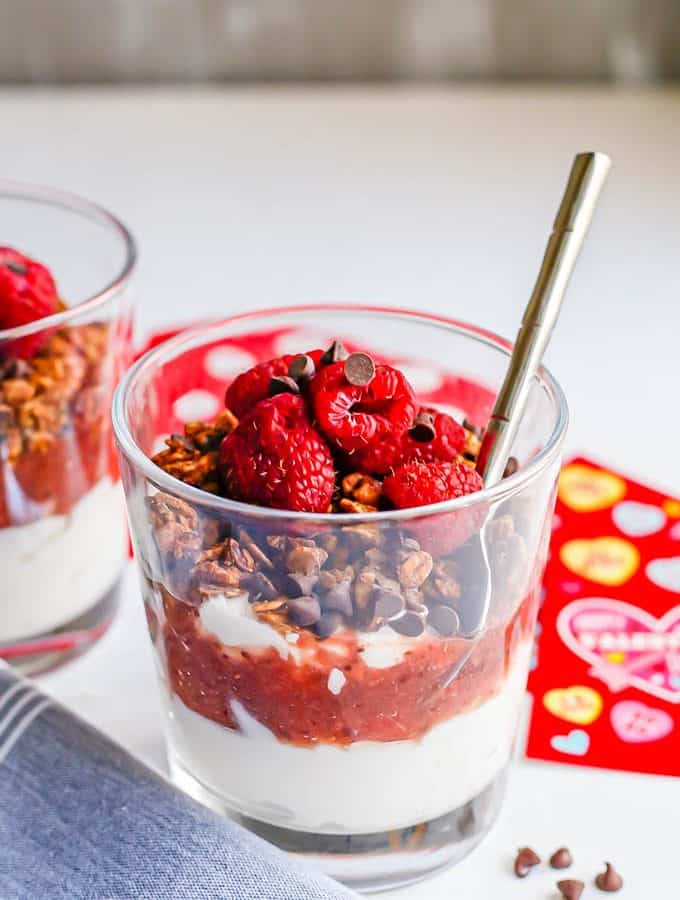 A layered yogurt parfait in a small clear glass with strawberry sauce, granola, berries and mini chocolate chips with a spoon tucked in the glass