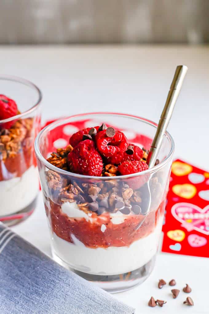 A layered yogurt parfait in a small clear glass with strawberry sauce, granola, berries and mini chocolate chips with a spoon tucked in the glass