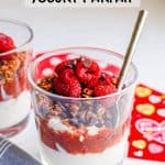 A layered yogurt parfait in a small clear glass with strawberry sauce, granola, berries and mini chocolate chips with a spoon tucked in the glass and a text overlay on the photo.