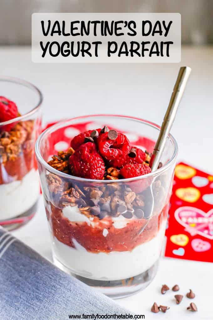 A layered yogurt parfait in a small clear glass with strawberry sauce, granola, berries and mini chocolate chips with a spoon tucked in the glass and a text overlay on the photo