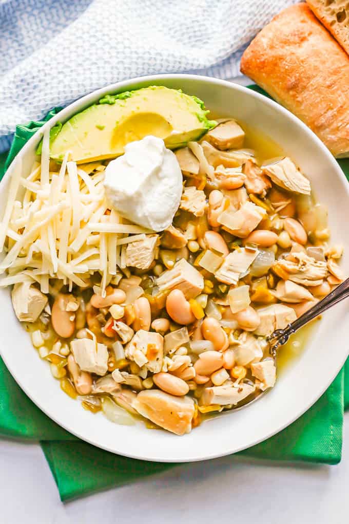 A white bowl with white chicken chili topped with cheese, sour cream and avocado and a spoon resting in the bowl