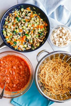 A spread of ingredients, including diced cooked chicken, cooked spaghetti noodles, a cheese and marinara sauce mixture and a skillet of sautéed vegetables