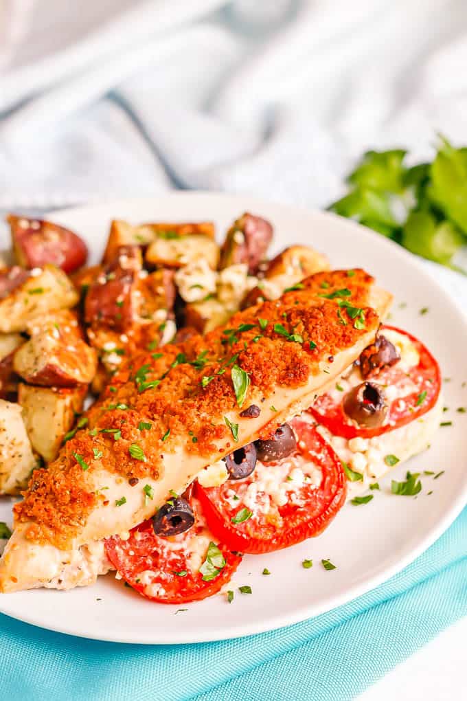 A stuffed chicken breast with tomatoes, olives and feta and a breadcrumb topping served on a white dinner plate with roasted potatoes