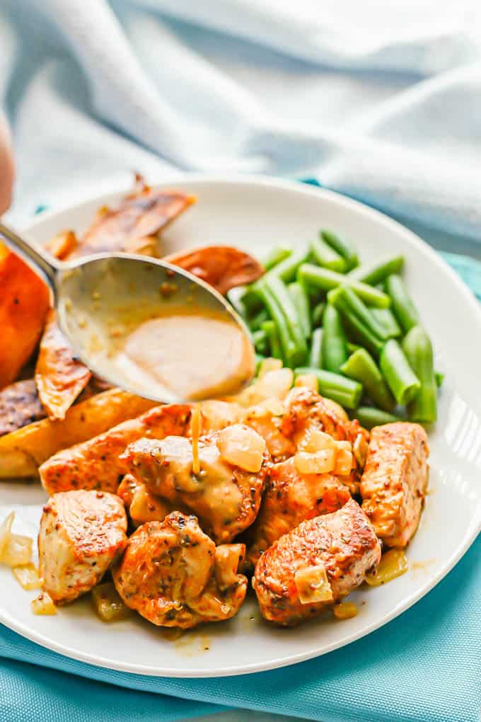 A spoon drizzling a honey mustard sauce over some cubed chicken pieces served on a white dinner plate