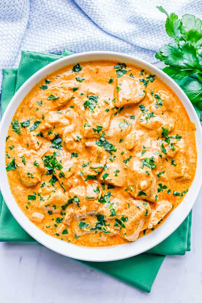 A white bowl full of butter chicken with chopped parsley on top and parsley sprigs to the side