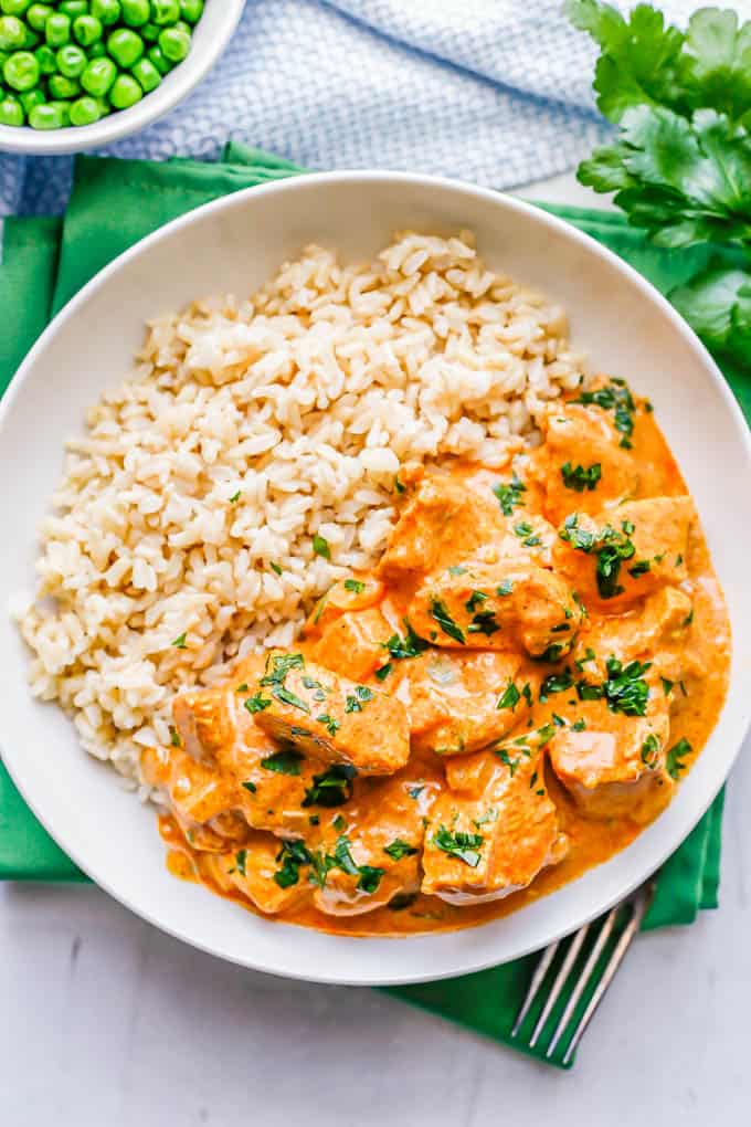 A low white bowl with Indian butter chicken alongside steamed rice with a bowl of peas to the side