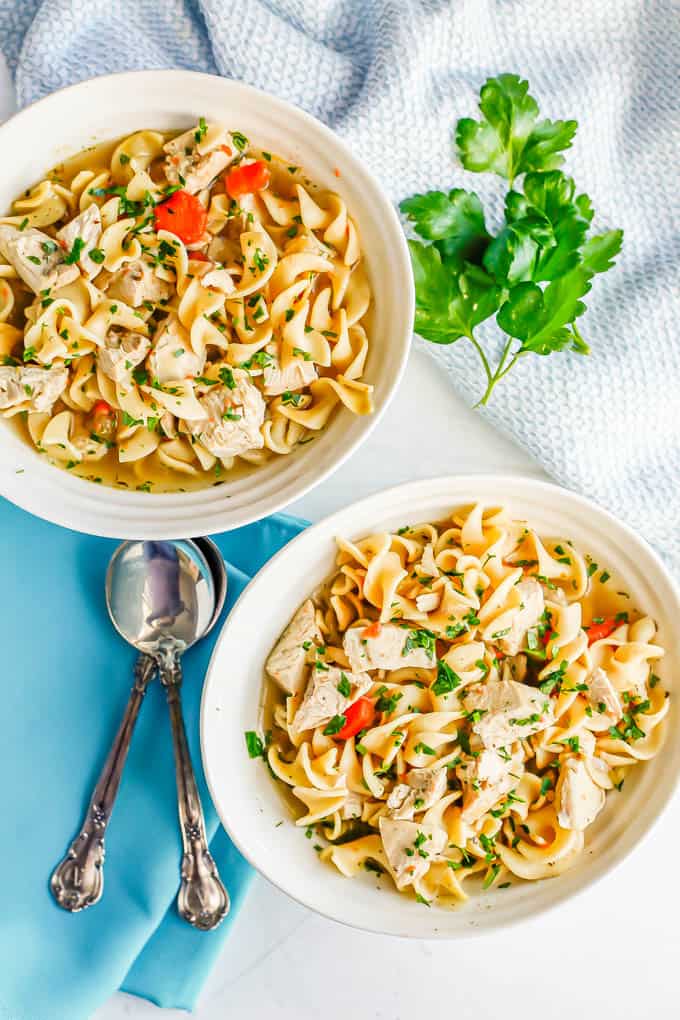 Two white bowls with servings of chicken noodle soup with spoons and sprigs of parsley to the side