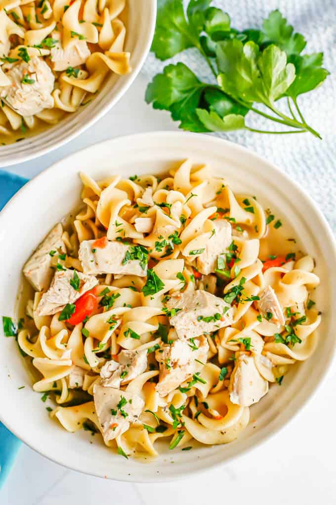 A white bowl full of chicken noodle soup with parsley sprinkled on top and sprigs of parsley to the side