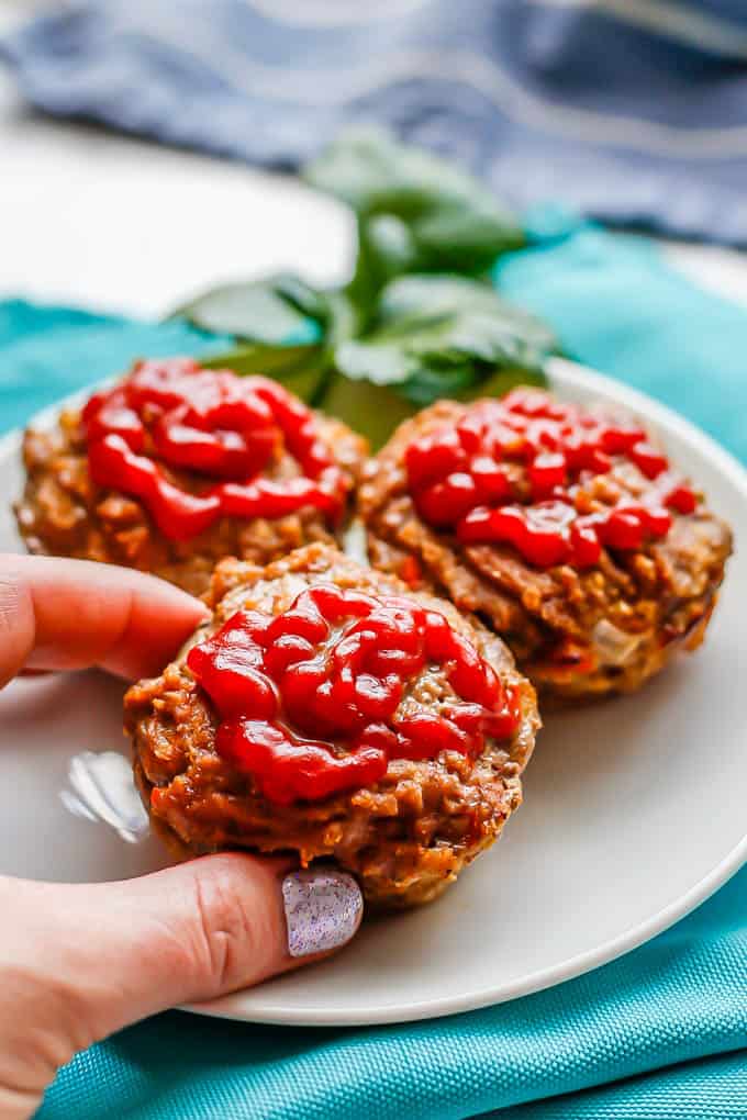 A hand picking up a mini meatloaf topped with ketchup from a white plate