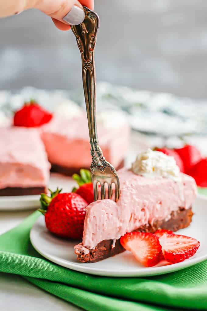 A hand holding a fork pressing into a slice of strawberry cheesecake with brownie crust served on a white plate