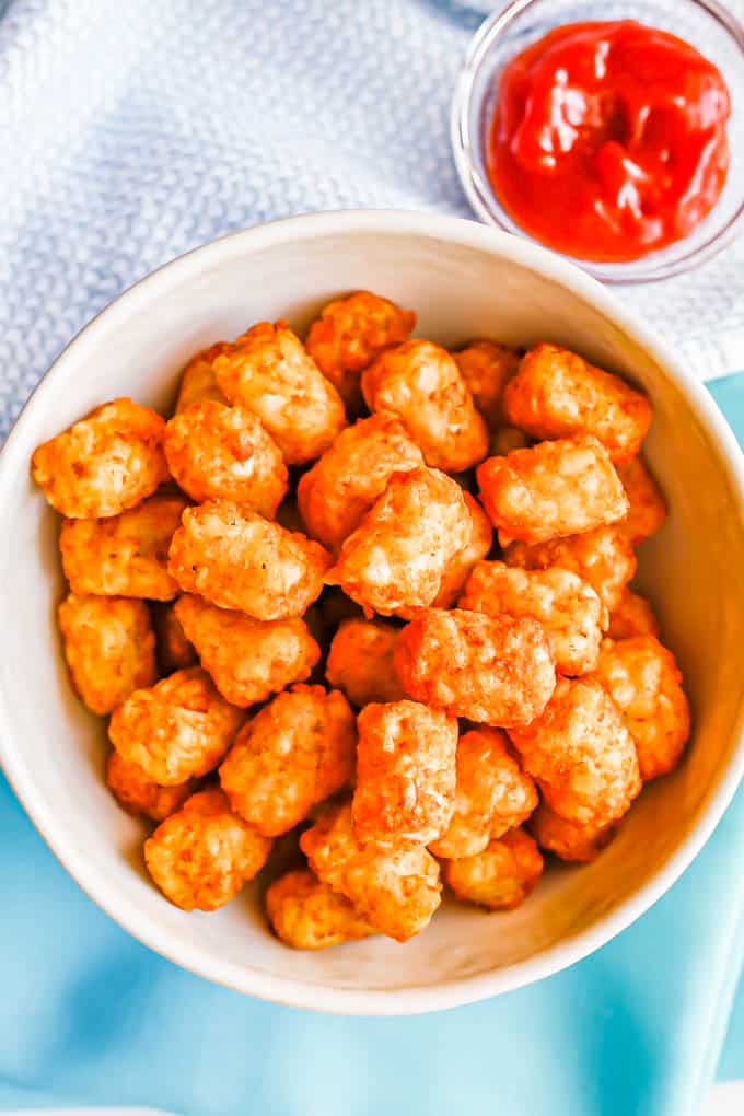 A white bowl of crispy tater tots served with a small bowl of ketchup to the side for dipping