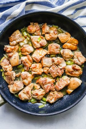 Seared chunks of chicken thighs being cooked in a large skillet with garlic and green onions