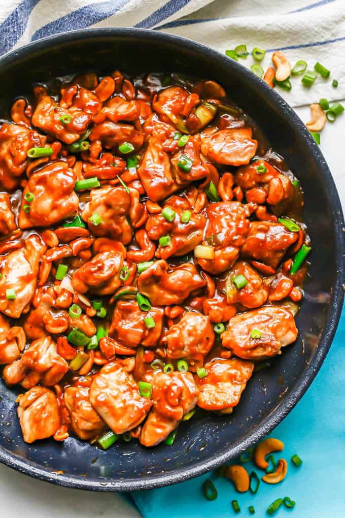 Cashew chicken with green onions in a large skillet with cashews and green onions to the side of the pan