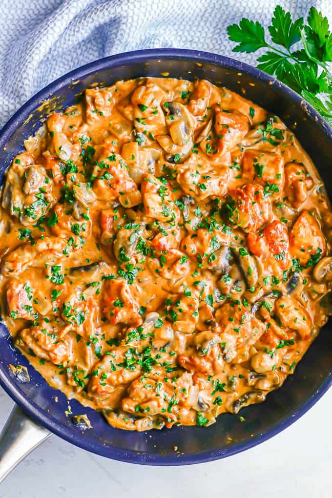 A large dark skillet with chicken paprikash in a creamy sauce with onions and mushrooms and topped with fresh chopped parsley