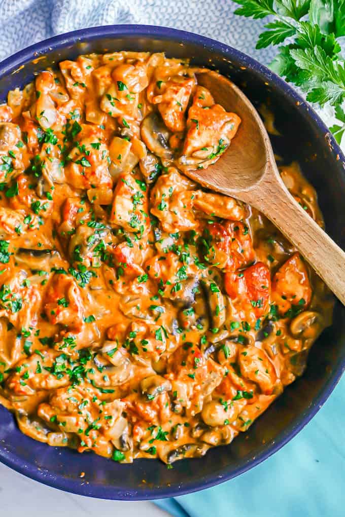 A wooden spoon resting in a large skillet with chicken paprika with onions and mushrooms