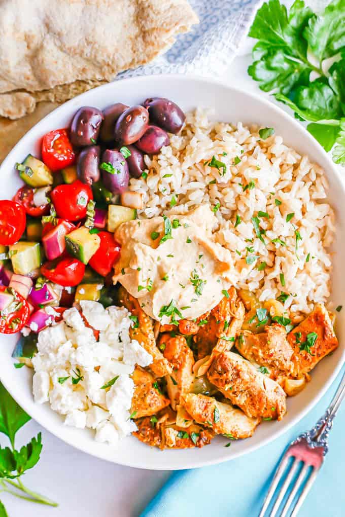 A low white bowl with brown rice, chicken shawarma, fresh veggies, olives, feta and hummus