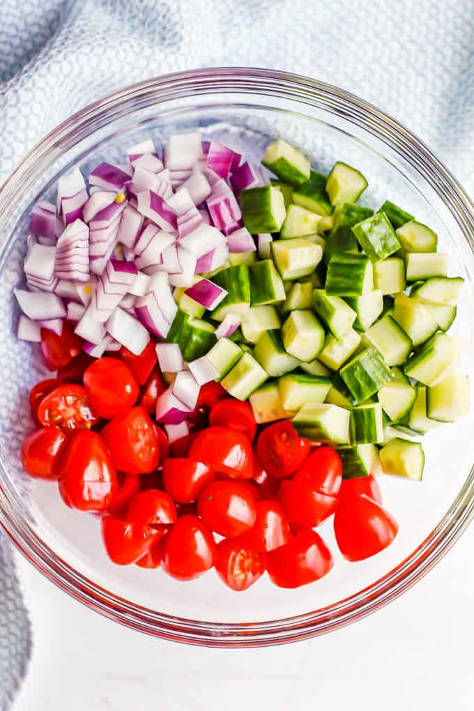 A clear bowl with chopped cucumbers, halved cherry tomatoes and chopped red onion arranged in piles