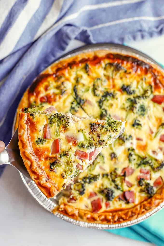A pie server lifting up a piece of baked quiche with ham, cheese and broccoli from a baking dish