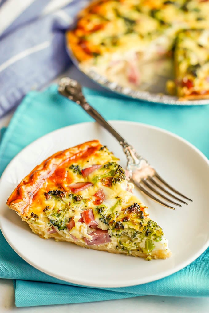 A slice of quiche with ham and broccoli served on a small white plate with a fork alongside and the pie plate of the baked quiche in the background