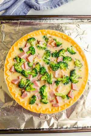 A pie shell filled with cheese, ham and broccoli and an egg mixture, set on a baking sheet before being cooked
