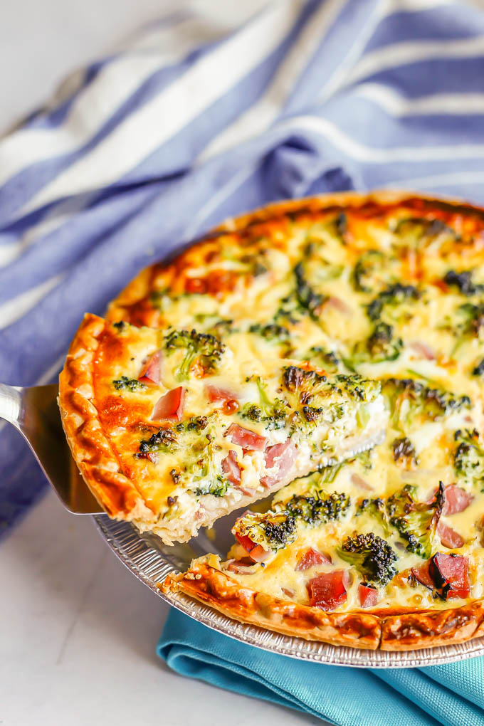 A pie spatula lifting up a slice of ham, egg and cheese quiche with broccoli from a baked dish