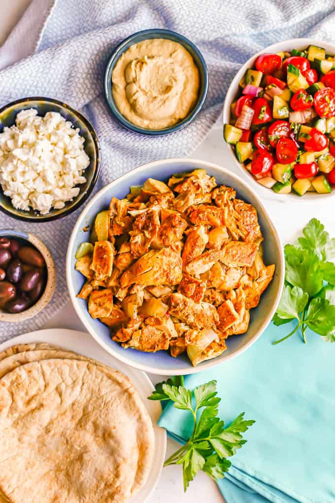 A bowl of cooked chicken shawarma surrounded by bowls of vegetables, hummus, feta cheese, olives and a plate of pita bread