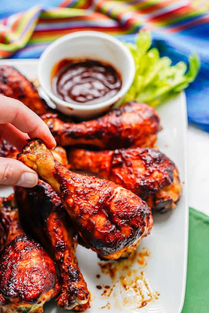 A hand picking up a BBQ chicken drumstick from a white platter