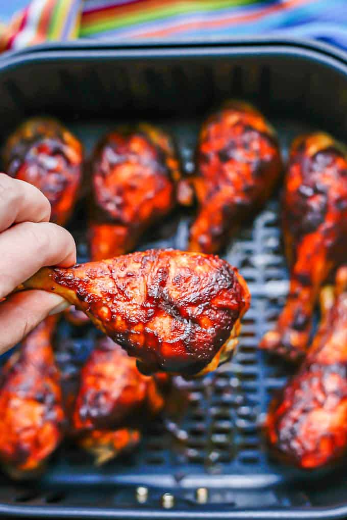 A hand holding up a cooked BBQ chicken leg from an Air Fryer tray full of chicken legs