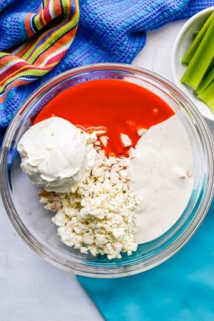 A large glass bowl with diced chicken, cream cheese, blue cheese crumbles, hot sauce and Ranch dressing