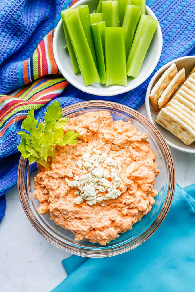 A buffalo chicken dip appetizer in a glass bowl with celery sticks and crackers in bowls nearby