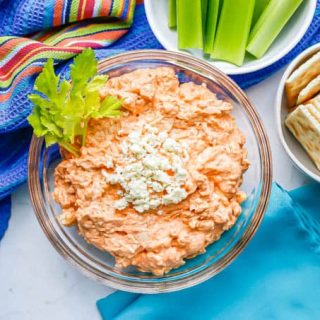 A glass bowl of a creamy chicken dip with blue cheese on top with celery and crackers nearby for dipping