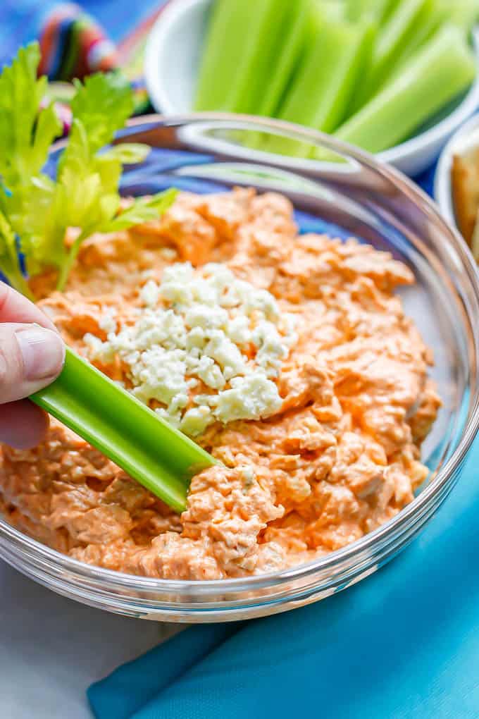 A hand dipping a celery stick into a glass bowl of buffalo chicken dip