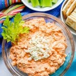 Buffalo chicken dip served in a glass bowl with blue cheese crumbles on top and a garnish of celery leaves with a text box on the photo