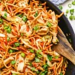 A wooden spoon resting in a large skillet of prepared chicken chow mein with a text overlay on the photo