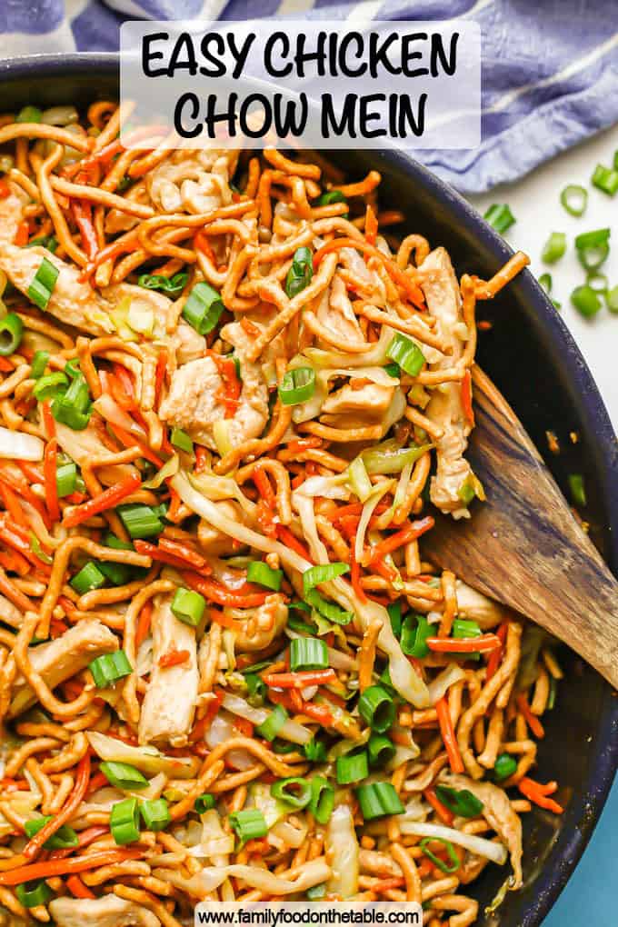 A wooden spoon resting in a large skillet of prepared chicken chow mein with a text overlay on the photo